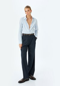 Camicia button-down azzurro chiaro con colletto aperto, abbinata a pantaloni sartoriali blu scuro e scarpe in pelle marrone, con una cintura marrone.