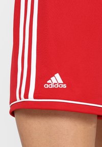 Røde atletikshorts med hvidt Adidas-logo og tre hvide lodrette striber langs siden, båret på en persons lår.