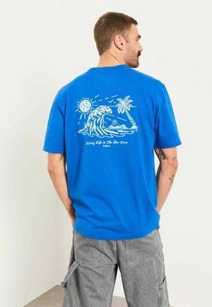 Mann trägt blaues T-Shirt mit weißem Motiv von Sonne, Welle, Palme und einer Person, die am Strand liegt, kombiniert mit grauer Cargo-Hose.