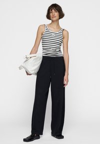 Gestreiftes Tanktop in Schwarz und Weiß, kombiniert mit locker sitzenden schwarzen Sweatpants. Das Model hält eine weiße Tasche mit gerolltem Rand und trägt schwarze Schuhe.