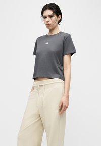 Νέος με κοντά σκούρα μαλλιά φοράει σκούρο γκρι κοντομάνικη μπλούζα και μπεζ φόρμα με κορδόνι, μπροστά σε απλό φόντο.