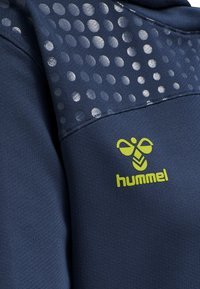 Top sportivo blu navy con spalla texturizzata, caratterizzato da un motivo di punti grigi chiari e un logo "hummel" verde brillante nella parte anteriore inferiore.