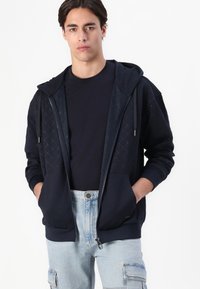 Felpa con zip di colore blu navy con pattern testurizzati sulla parte anteriore, dotata di cappuccio e due tasche laterali, abbinata a pantaloni in denim chiaro.