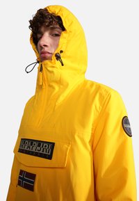Yellow waterproof jacket with a high collar, adjustable hood, front pocket, and branded patches. Smooth texture and minimalist design.