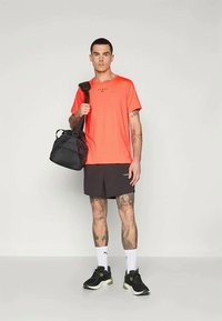 Férfi élénk narancssárga pólóban és fekete rövidnadrágban, fekete edzőtáskát tart a vállán, fehér zoknit és fekete sportcipőt visel, sima háttér előtt áll.