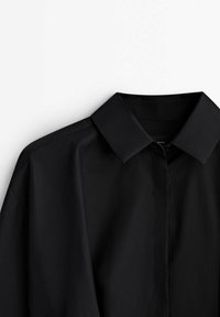 Schwarzes Langarmshirt mit spitzem Kragen, glatter Textur, subtiler Glanz und durchgehender Knopfleiste. Sauberes, minimalistisches Design.