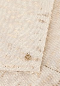 Beiger Textilstoff mit metallischen goldenen Fäden, ausgestattet mit einem kleinen runden goldenen Emblem an der Faltung. Glatte und elegante Oberfläche.