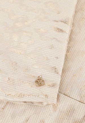 Beige gestructureerde stof met metallic gouden draadjes, met een klein circulair gouden embleem bij de vouw. Glad en verfijnd afwerking.