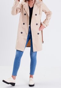 Beige trenchcoat met een dubbelknopen ontwerp, grote zwarte knopen en een trekkoordcapuchon. Gecombineerd met blauwe skinny jeans en witte schoenen.