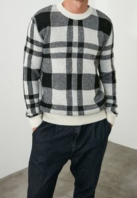 Suéter a cuadros en blanco y negro con cuello redondo, puños acanalados y un corte relajado. Lleva encima pantalones oscuros sobre un fondo liso.