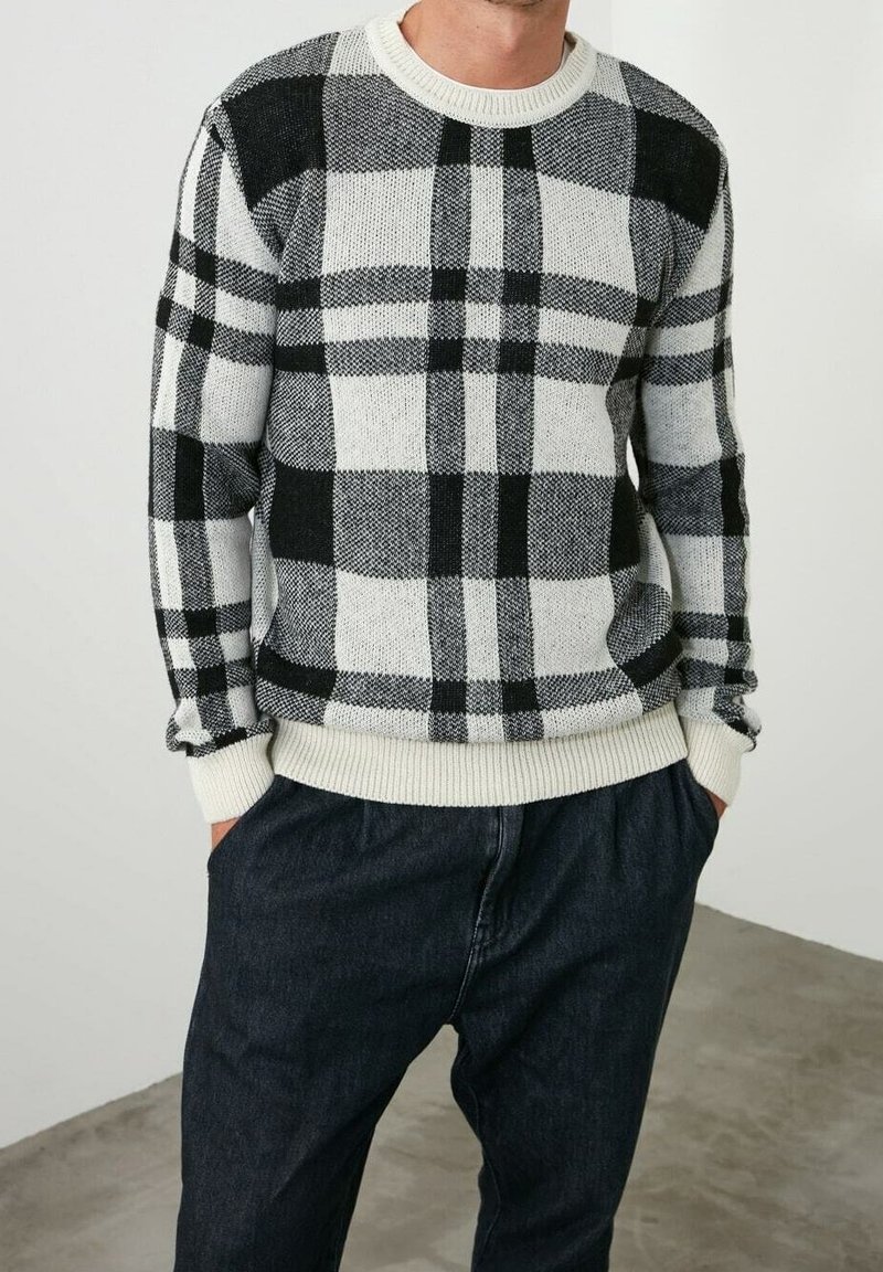 Suéter a cuadros en blanco y negro con cuello redondo, puños acanalados y un corte relajado. Lleva encima pantalones oscuros sobre un fondo liso.