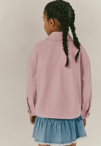 Veste en velours côtelé rose rayé avec des boutons, portée sur une jupe en denim bleu clair à volants. Tissu doux avec une coupe décontractée.