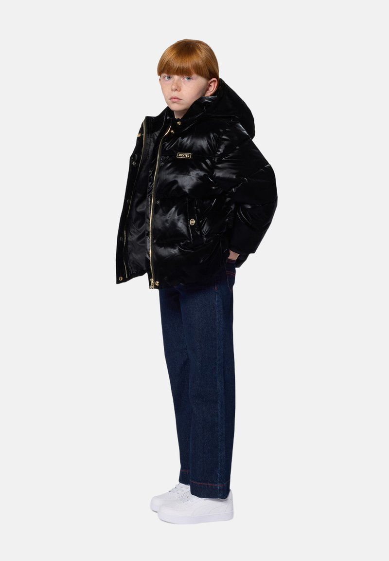Enfant aux cheveux roux portant une doudoune noire brillante, un jean bleu foncé et des baskets blanches, se tenant devant un fond uni.