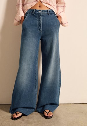 Weite blaue Denim-Jeans mit einem ausgewaschenen Finish, Knopfverschluss und Fronttaschen, kombiniert mit schwarzen Flip-Flops. Angenehme Textur und lässige Passform.