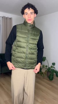 Νεαρός με σγουρά μαλλιά, φορεμένο πράσινο μακρύ γιλέκο κεντημένο πάνω από μαύρο μακρυμάνικο πουκάμισο και μπεζ παντελόνια, στέκεται μέσα σε εσωτερικό χώρο κοντά σε φυτά.