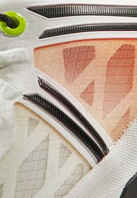 Buty z białym, teksturowanym materiałem wierzchnim, ozdobione pomarańczowymi i beżowymi wzorami geometrycznymi, kontrastującymi czarnymi akcentami oraz zielonymi detalami w postaci oczek.