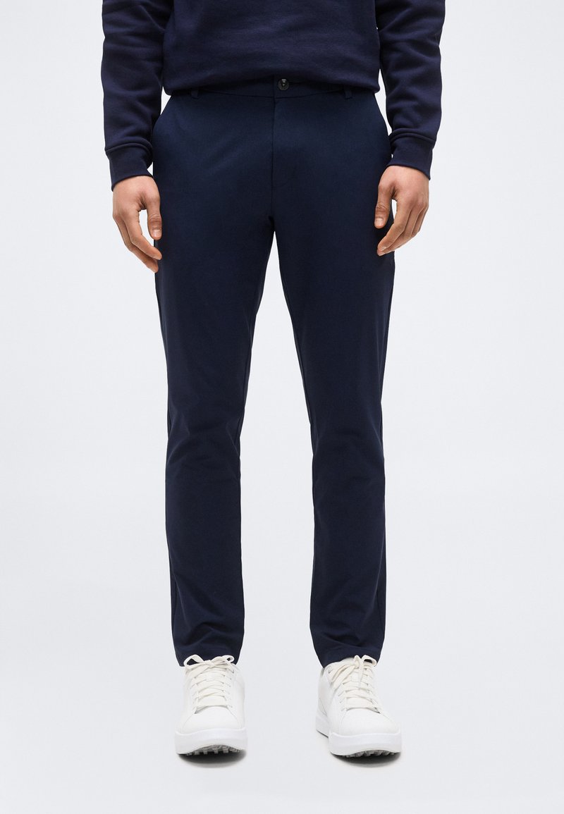 Homme portant un pantalon slim bleu marine et des baskets blanches, debout devant un fond uni clair.