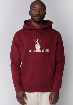 Man draagt een bordeauxrode hoodie met een afbeelding van gekruiste vingers en de tekst "Fingers Crossed" aan de voorkant tegen een effen achtergrond.