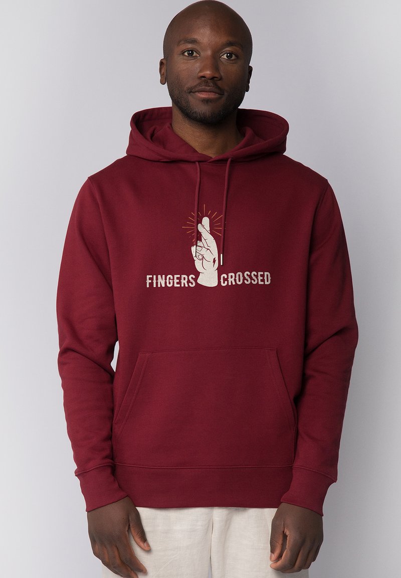 Homme portant un sweat à capuche bordeaux avec un dessin de doigts croisés et le texte "Fingers Crossed" sur le devant, sur un fond uni.