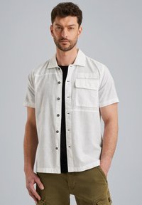 Camisa blanca ligera de manga corta con bot�ón en el frente, un bolsillo en el pecho y un acabado ligeramente texturado. Llevada sobre una camiseta negra.