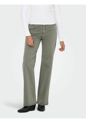 Donna che indossa jeans verde oliva a gamba dritta, stivali neri con tacco e una maglia bianca a maniche lunghe su uno sfondo chiaro e uniforme.