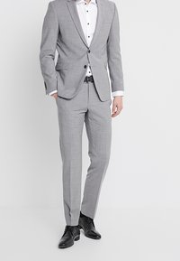 Ljusgrå kostym med ett texturerat mönster, kombinerad med en vit skjorta med svarta knappar. Utseendet fulländas med svarta dresskor.