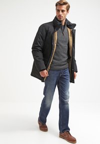 Chaqueta aislante negra y marrón con un cuello alto, suéter de punto gris debajo, combinada con jeans azules y zapatos marrones. Diseño sencillo.