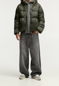 Chaqueta hinchada verde oliva con capucha, combinada con una sudadera gris y vaqueros grises sueltos y desgastados. Zapatillas grises y blancas completan el atuendo.