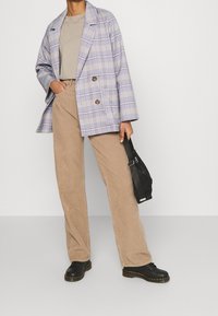 Personne portant un pantalon en velours côtelé beige, un t-shirt gris, un blazer à carreaux violet clair, des chaussures noires, tenant un petit sac noir, debout devant un fond uni.