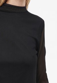 Close-up van een vrouw die een zwarte top met hoge hals draagt, met doorzichtige lange mouwen en een getextureerde stof.