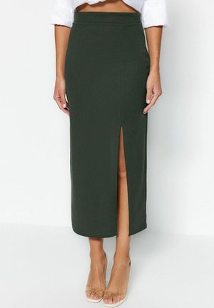 Dunkelgrüner Midi-Bleistiftrock mit Frontschlitz, kombiniert mit weißem Top und transparenten High-Heel-Schuhen vor einfachem Hintergrund.