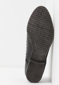 Semelle de chaussure noire en caoutchouc texturé pour l'adhérence, présentant un motif répété du logo "Tamaris" et un indicateur de taille "37".