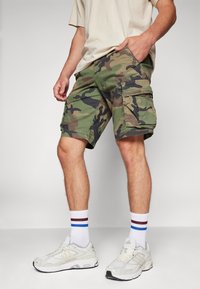 Camo cargo shorts in green, brown, and black with multiple pockets. Worn with white sneakers and striped crew socks. Casual fit.