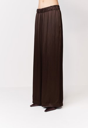 Tmavohnedé široké nohavice s elastickým pásom, nosené s topánkami so špicatou špičkou na svetlom pozadí.