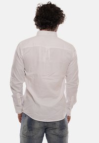 Camicia bianca a maniche lunghe con vestibilità rilassata, colletto a bottone e dettaglio sul yoke posteriore, abbinata a jeans blu sbiaditi.