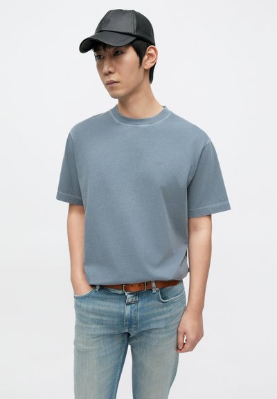 Μπλε βαμβακερό t-shirt με κοντά μανίκια και στρογγυλή λαιμόκοψη, σε συνδυασμό με ανοιχτόχρωμο μπλε τζιν παντελόνι και μαύρο καπέλο, με καφέ ζώνη.