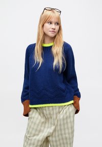 Suéter de punto azul marino con puños en verde neón y marrón, con un patrón en zigzag a lo largo del dobladillo. Combinado con pantalones a cuadros claros.