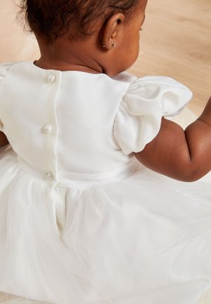 Jeune enfant portant une robe blanche avec des manches bouffantes et des boutons en perles, assis sur un sol en bois clair, vu de dos.