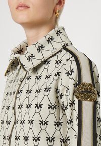 Chaqueta beige con patrón floral negro, cremallera dorada y cuello ancho. Presenta un parche texturizado en el labio y franjas laterales con acentos dorados y negros.