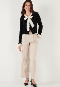 Black cardigan with lace trim and bow detail, paired with beige wide-leg trousers. The outfit is complemented by black heeled shoes.
