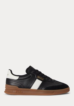 Černý kožený teniska s bílým bočním pruhem a detailem na patě, hnědá gumová podrážka a logo "Polo" poblíž tkaniček a podrážky.