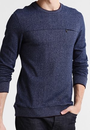 Man draagt een nauwsluitende donkerblauwe trui met lange mouwen en een klein ritsvakje op de borst, hand in de zak, tegen een effen achtergrond.