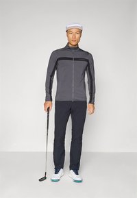 Gray athletic jacket with black horizontal stripe, paired with dark pants and white shoes. Model holds a golf club, wearing a white cap.