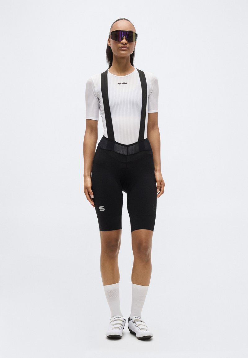 Γυναίκα που φορά λευκή ποδηλατική μπλούζα με κοντά μανίκια, μαύρο σορτς με τιράντες, λευκές κάλτσες, λευκά ποδηλατικά παπούτσια και γυαλιά ηλίου με ανακλαστική επίστρωση.