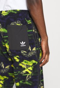 Adidas fleecebyxor i svart, grön och gul tie-dye-mönster. Har en svart sidoficka och tre svarta ränder längs benet.