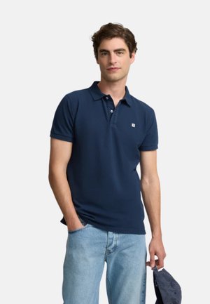 Polo de color azul marino con cuello, mangas cortas y un pequeño logo en el pecho. Llevado con unos jeans de color azul claro, textura de tela suave.