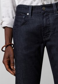 Mörka denimjeans med rak passform, diskreta sömmar, två framfickor och en bakficka. Bärs med en uppvikt vit skjortärme.