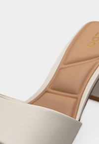 Gros plan d'une sandale à talon beige avec semelle intérieure rembourrée couleur fauve et bout ouvert sur fond blanc.