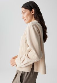 Beige corduroy overhemdjasje met een enkele voorzak, een relaxte pasvorm en lange mouwen. Het heeft een verticale ribbelstructuur en subtiele openingen aan de onderkant.