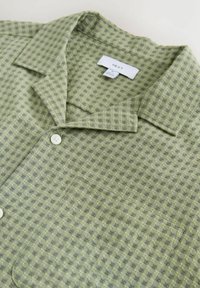 Zaļš un pelēks raibuma struktūras krekls ar iegriezto apkakli, priekšējo kabatu un baltiem pogām, izlikts plakani ar redzamu zīmola etiķeti.